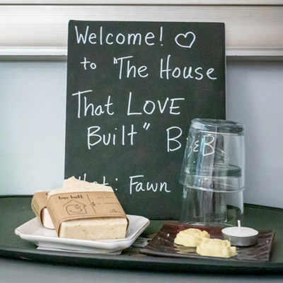 Soap, some cookies and a glass sit on top of a table in front of a sign that reads "Welcome to The House That Live Built Bed and Breakfast.